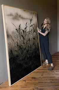 Asheville Artist Linsey Gray standing beside and admiring her large charcoal and soft pastel on wood artwork, titled "Breathe Now", featuring moody silhouettes of oversized wild grasses with faint mountain views in the distance. 