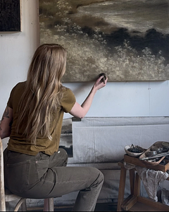 Asheville artist, Linsey Gray, working in her Asheville studio on her charcoal and soft pastel on wood piece, titled "Silent Echo", featuring an abstract shoreline and a palette of blacks, cool grays, earthy greens, browns, and golds.