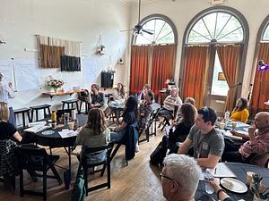 This photo includes the interior of Story Parlor with large art deco windows in the background. Seated in the foreground are people of multiple demographics engaging in conversation at round tables.