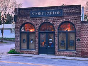 This image includes an angled view of Story Parlor's exterior at sunset. It is an art deco era brick building with half crescent high windows over long rectangular windows. The doors are center of the building. There is a sign above the doors that reads "Story Parlor".